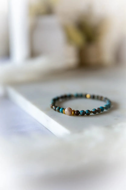 Brązowo-niebieska bransoletka z agatów i turkusów z jasnoróżowym kamieniem centralnym i złotymi przekładkami na jasnym tle.