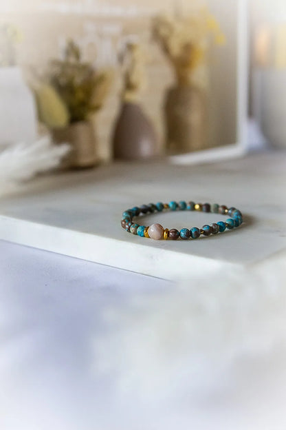 Bransoletka 'Blekitna Rownowaga' z turkusowych i brązowych agatów, z okrągłym, różowym kamieniem pośrodku. Ułożona na jasnym tle z delikatnie rozmytym, dekoracyjnym tłem.