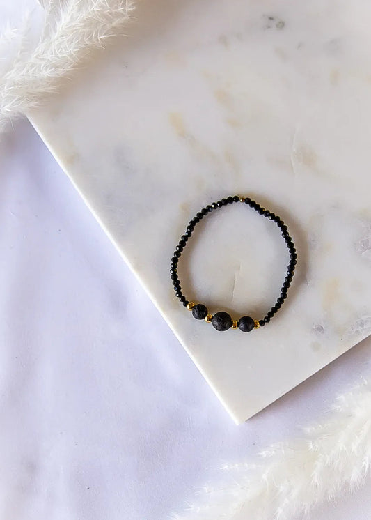 Czarna Moc Bransoletka Z Czarnej Lawy Turmalin Hematyt: subtelna, czarna bransoletka z lawy i ozdobnymi złotymi przekładkami na marmurowym tle, otulona puszystą trawą.