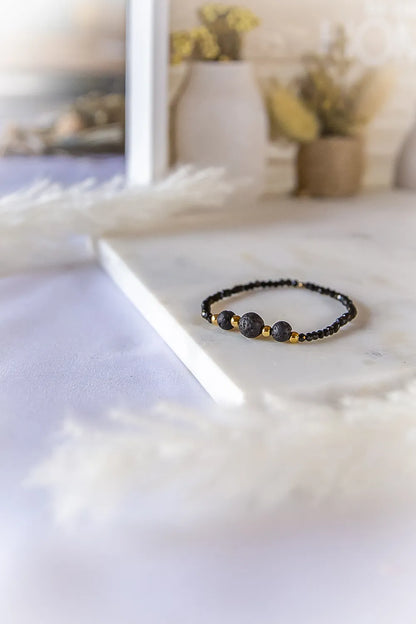 Czarna Moc Bransoletka Z Czarnej Lawy Turmalin Hematyt - skupina 3 czarnych kamieni lawy i złotych kulek na tle białego marmuru.