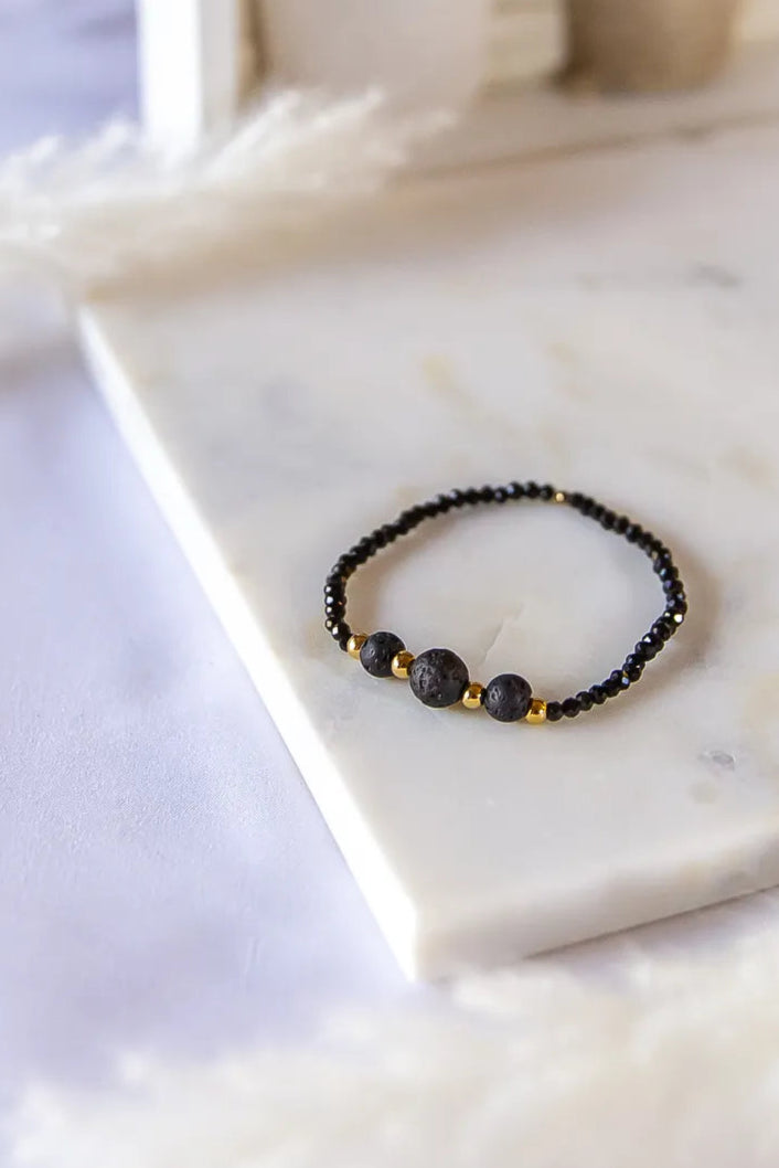 Czarna Moc Bransoletka Z Czarnej Lawy Turmalin Hematyt: closeup matowej czarnej bransoletki z trzema okrągłymi kamieniami lawy, ozdobionej złotymi hematytami, ułożonej na marmurowej płycie na tle białej, puszystej trawy.