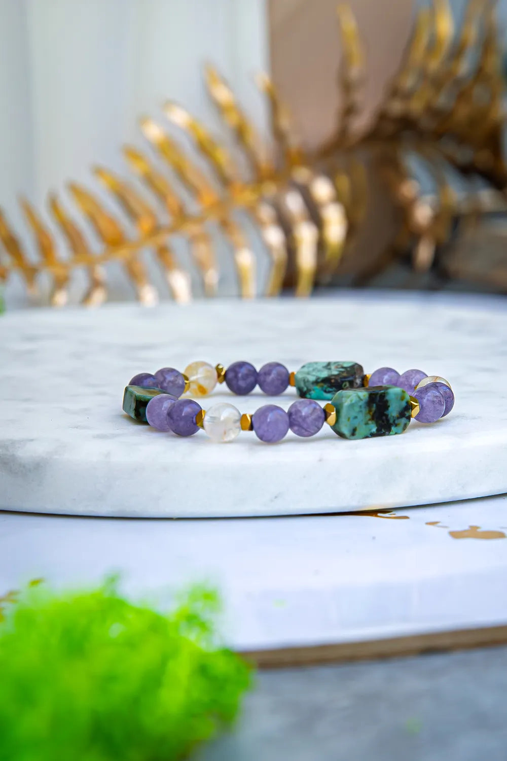 Bransoletka 'DROGA DUSZY' z turkusem afrykańskim, ametystem i cytrynem, ułożona na białym marmurowym tle z zielonymi liśćmi w tle.