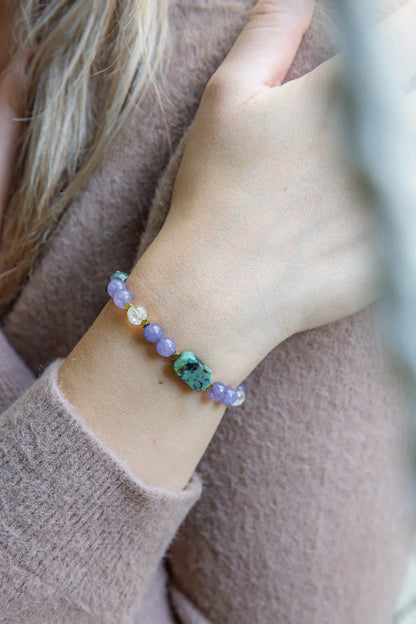 Bransoletka 'DROGA DUSZY' z turkusem afrykańskim, ametystem i cytrynem, widoczna na nadgarstku kobiety. Kamienie ułożone w szereg na delikatnym tle.