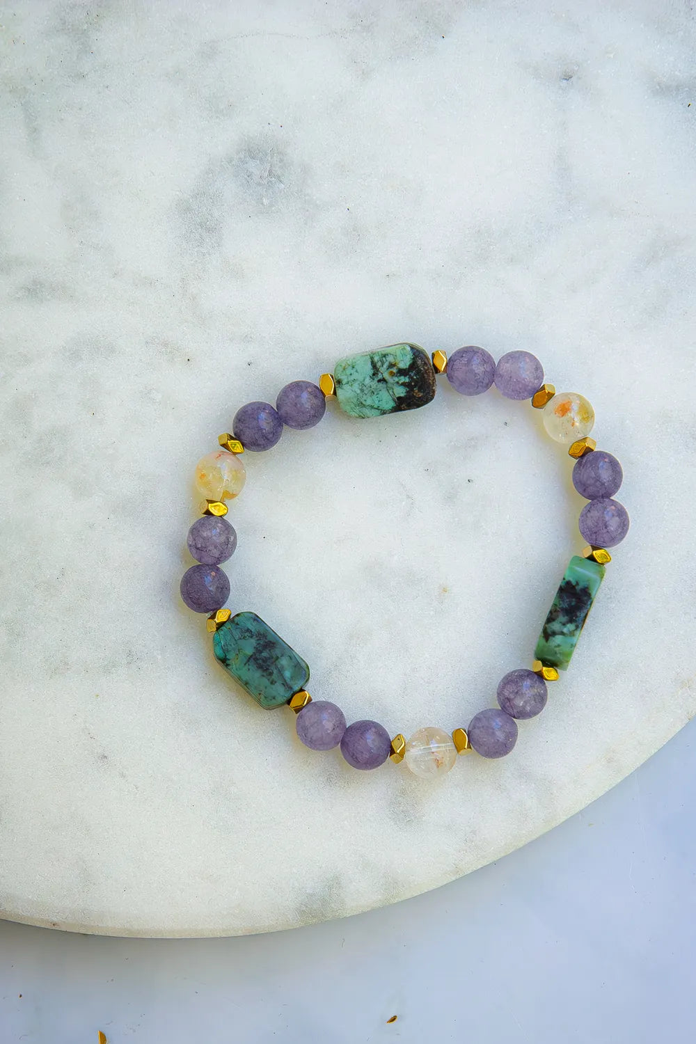 DROGA DUSZY - BRANSOLETKA Z TURKUSU AFRYKAŃSKIEGO, AMETYSTU I CYTRYNU. Bransoletka z kolorowymi kamieniami, w tym turkus afrykański, ametyst i cytryn, ułożona na białym marmurowym tle.