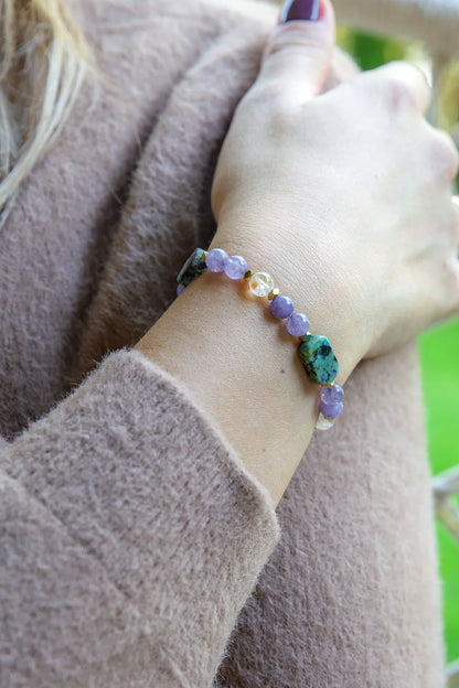 DROGA DUSZY - BRANSOLETKA Z TURKUSU AFRYKAŃSKIEGO, AMETYSTU I CYTRYNU. Bransoletka z kolorowymi kamieniami, w tym turkusowym, fioletowym i żółtym, na tle delikatnej, beżowej bluzy. Ręka trzyma się ramienia, ułożenie naturalne.