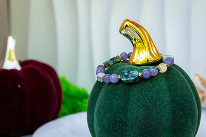 DROGA DUSZY - BRANSOLETKA Z TURKUSU AFRYKAŃSKIEGO, AMETYSTU I CYTRYNU. Bransoletka z kolorowymi kamieniami, w tym turkus, ametyst i cytryn, umieszczona na zielonym aksamitnym dyniowym podstawce. Tło z czerwonymi i zielonymi dekoracjami.