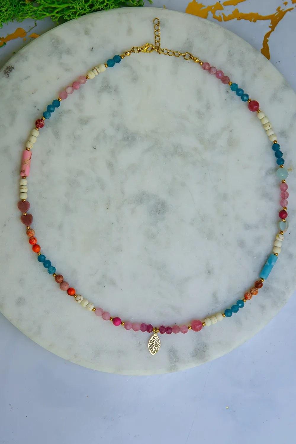 FESTIWAL RADOŚCI - NASZYJNIK Z JADEITU TURMALINOWEGO, TURMALINU I JASPISU. Naszyjnik z kolorowymi koralikami jadeitu turmalinowego, turmalinu i jaspisu, ułożony na marmurowym tle z zielonymi liśćmi w tle.