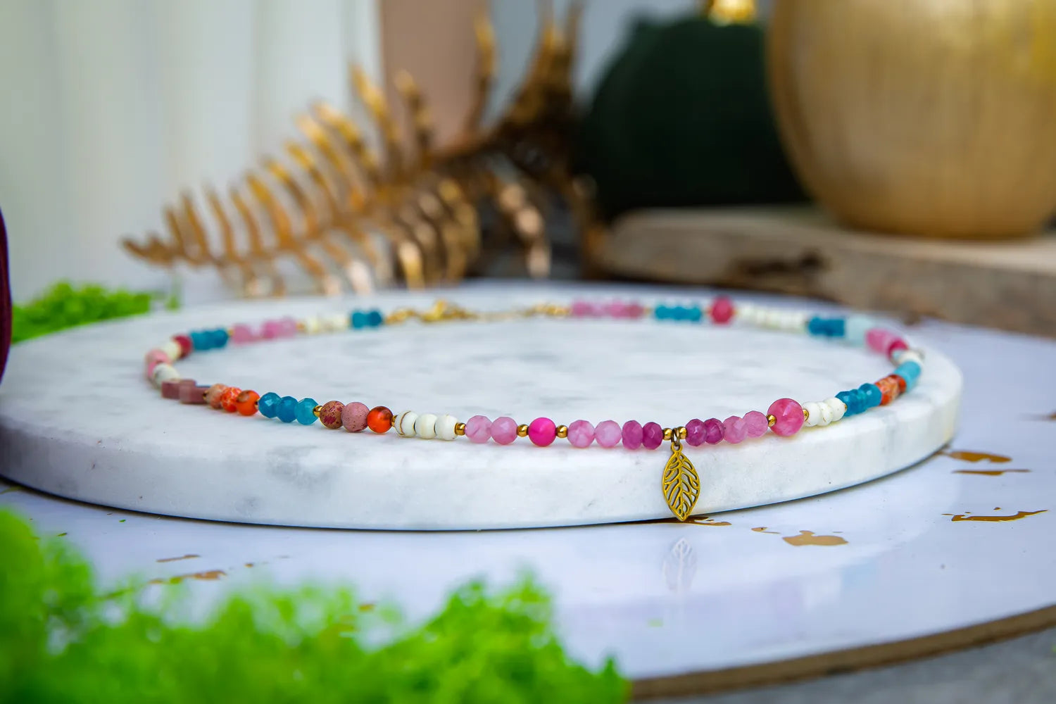 FESTIWAL RADOŚCI - NASZYJNIK Z JADEITU TURMALINOWEGO, TURMALINU I JASPISU. Naszyjnik z kolorowymi kamieniami, w tym jasnozielonymi, różowymi i pomarańczowymi, na białym marmurowym tle z dekoracją zielonych liści i złotych elementów.