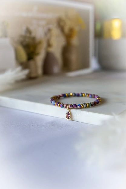 Fioletowa Zorza Bransoletka Z Jaspisu Kamienie z pojedynczym, owalnym kamieniem w złotym etui, ułożona na białej powierzchni, obok niewyraźnych dekoracji.