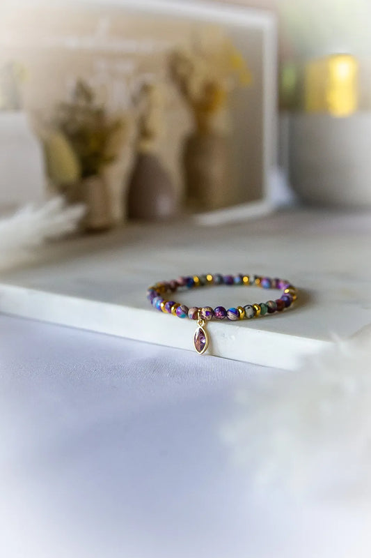 Fioletowa Zorza Bransoletka Z Jaspisu Kamienie z pojedynczym, owalnym kamieniem w złotym etui, ułożona na białej powierzchni, obok niewyraźnych dekoracji.