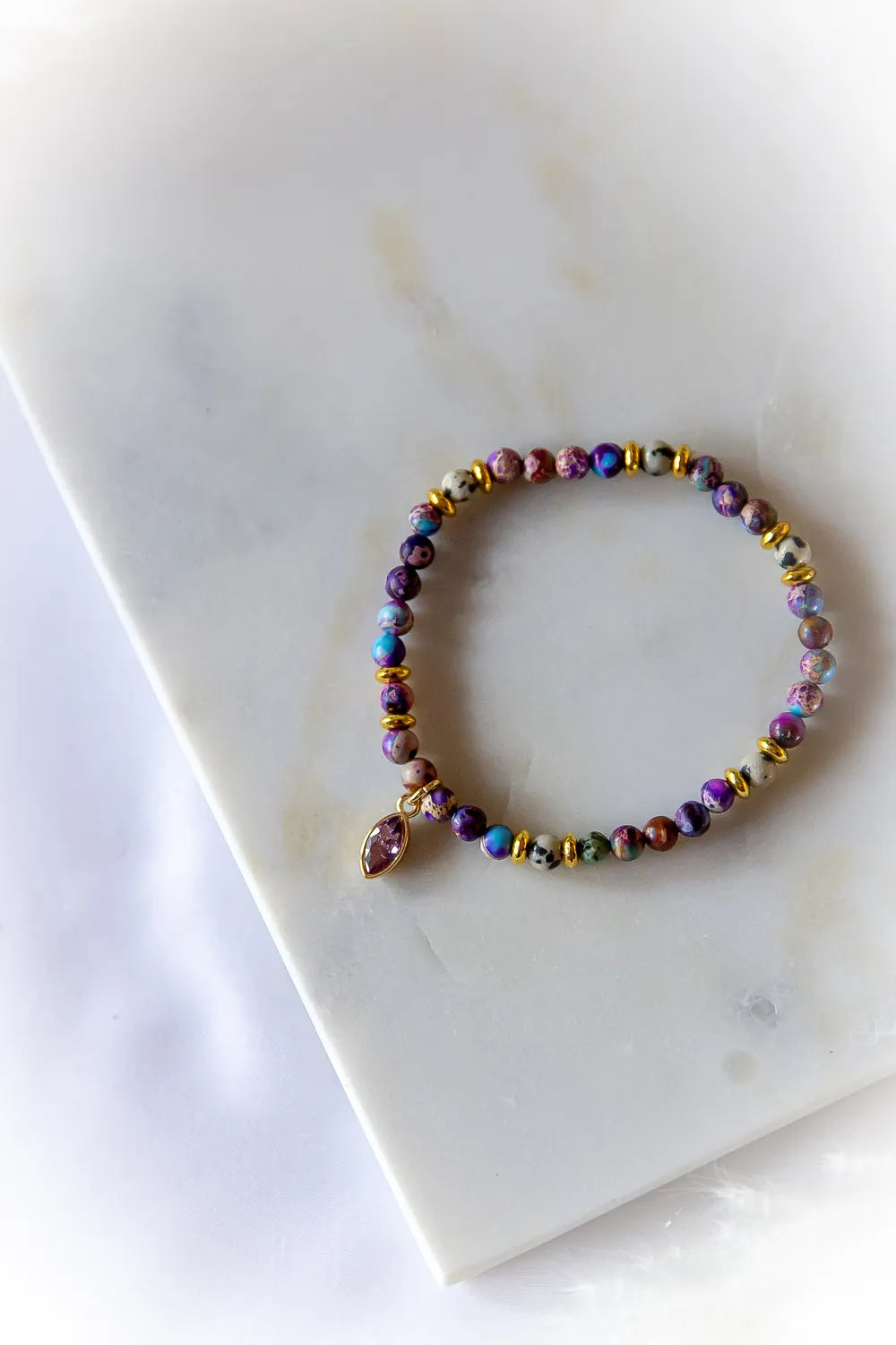 Fioletowa Zorza Bransoletka Z Jaspisu Kolorowe Koraliki, z błyszczącym złotym przekładkami i wiszącym kamieniem, na tle z białego marmuru.