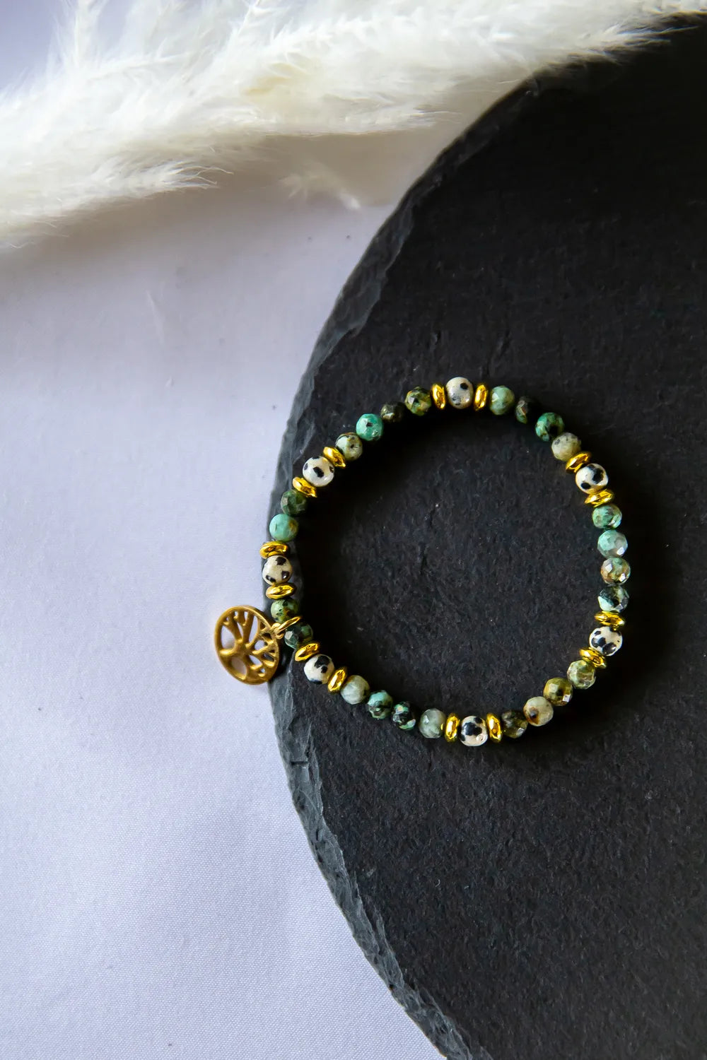 Gaj Madrosci Bransoletka Z Turkusem I Jaspisem, prezentowana na czarnym kamieniu, z zielonymi i białymi kamieniami oraz złotymi przekładkami. Urokliwe boho detale.