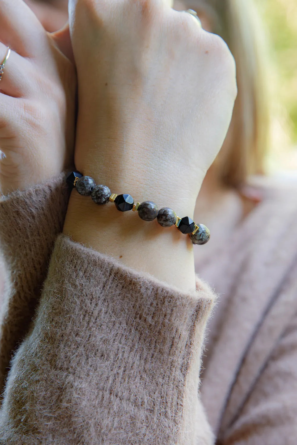 GŁĘBOKIE KORZENIE - BRANSOLETKA Z BRĄZOWEGO OBSYDIANU I ONYKSU na nadgarstku, z widocznymi okrągłymi kamieniami i złotymi elementami, na tle delikatnej, beżowej bluzy.
