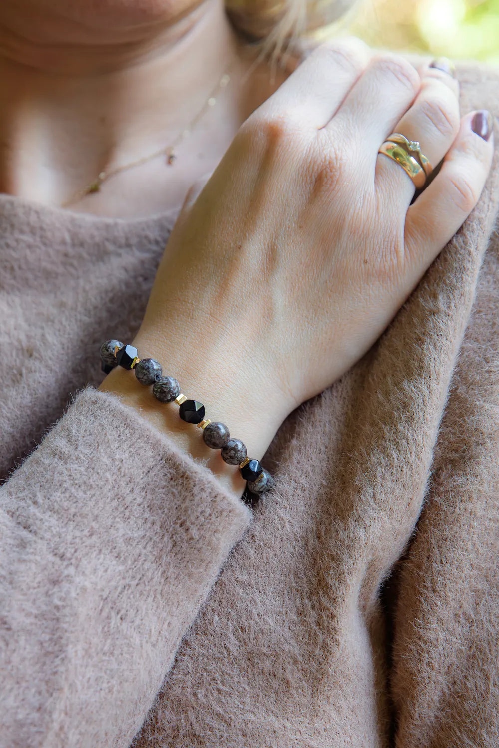GŁĘBOKIE KORZENIE - BRANSOLETKA Z BRĄZOWEGO OBSYDIANU I ONYKSU na nadgarstku, z widocznymi brązowymi i czarnymi kamieniami, na tle beżowego swetra.