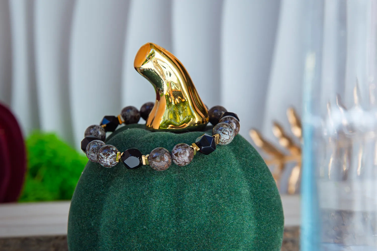 GŁĘBOKIE KORZENIE - BRANSOLETKA Z BRĄZOWEGO OBSYDIANU I ONYKSU, prezentowana na zielonym podstawce z złotym akcentem, z widocznymi kamieniami brązowego obsydianu i czarnego onyksu, na tle białych zasłon i dekoracji.