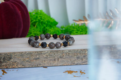 GŁĘBOKIE KORZENIE - BRANSOLETKA Z BRĄZOWEGO OBSYDIANU I ONYKSU, prezentująca się na drewnianym tle z zielonym mchem w tle. Biżuteria składa się z brązowych kulek obsydianu i czarnych kulek onyksu, ułożonych w elegancki wzór.