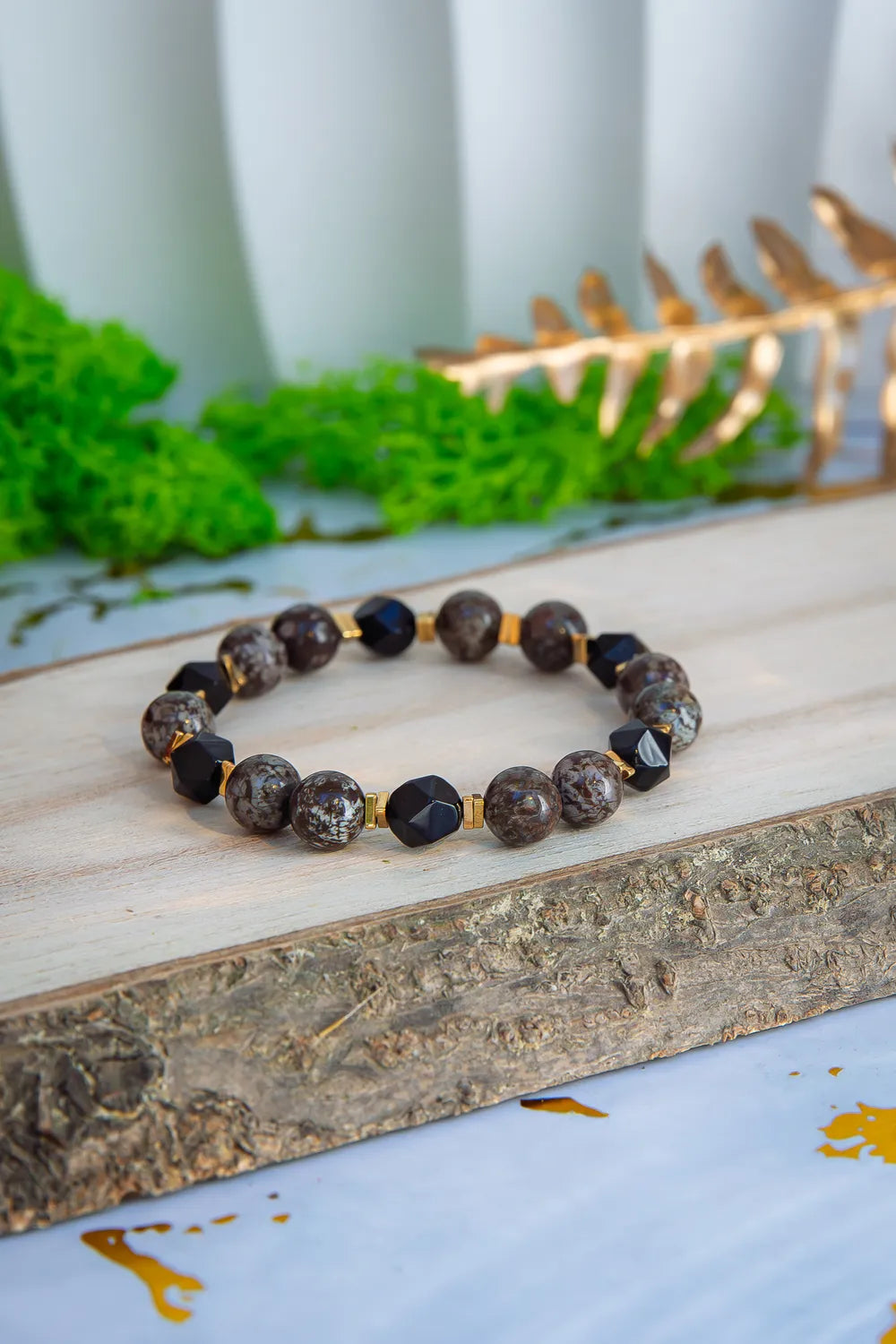 GŁĘBOKIE KORZENIE - BRANSOLETKA Z BRĄZOWEGO OBSYDIANU I ONYKSU, prezentująca się na drewnianym tle z zielonym mchem i złotymi liśćmi w tle. Biżuteria składa się z okrągłych kamieni brązowego obsydianu i czarnych kryształów onyksu.