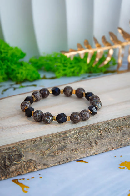 GŁĘBOKIE KORZENIE - BRANSOLETKA Z BRĄZOWEGO OBSYDIANU I ONYKSU, prezentująca się na drewnianym tle z zielonym mchem i złotymi liśćmi w tle. Biżuteria składa się z okrągłych kamieni brązowego obsydianu i czarnych kryształów onyksu.