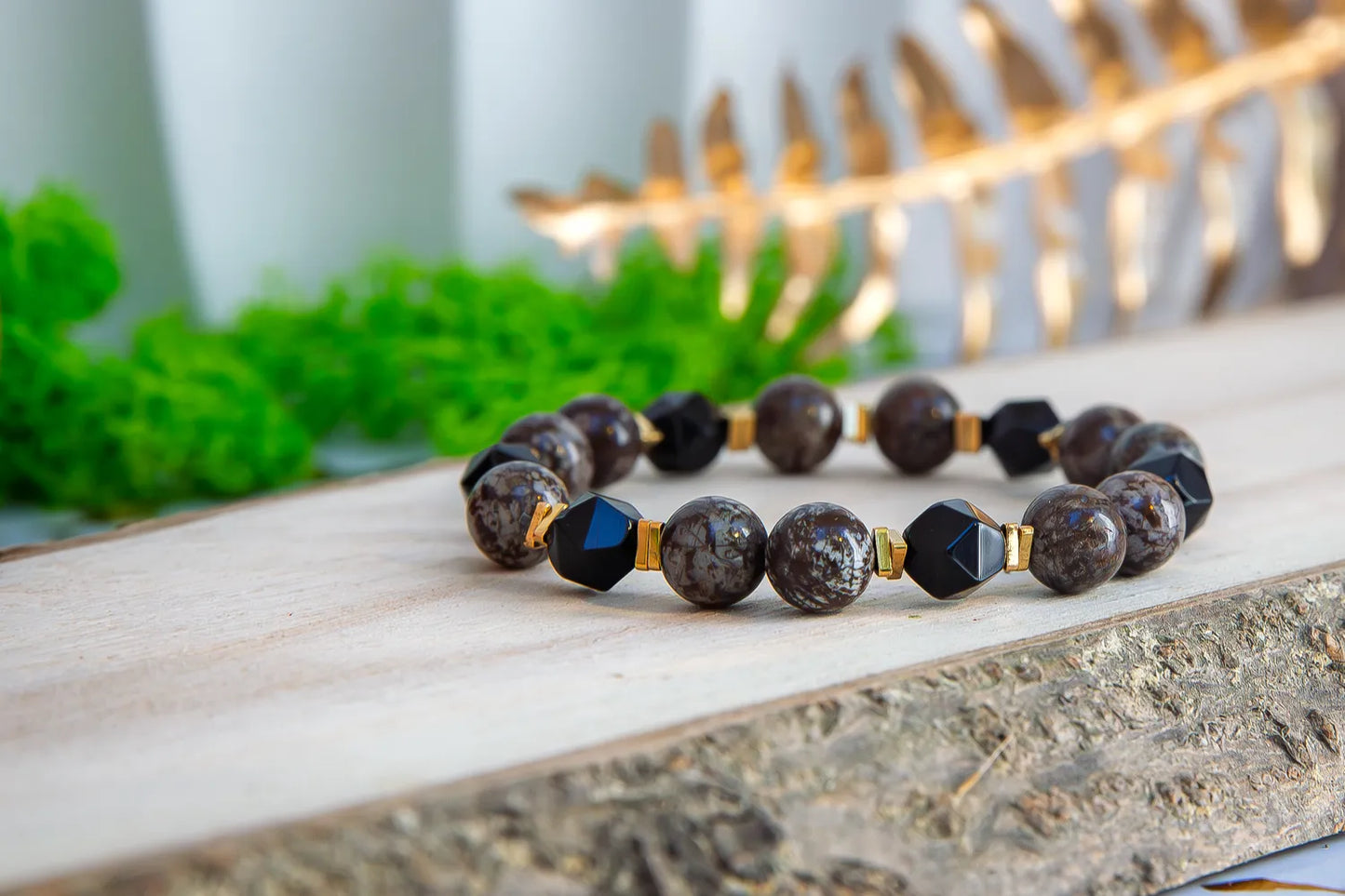 GŁĘBOKIE KORZENIE - BRANSOLETKA Z BRĄZOWEGO OBSYDIANU I ONYKSU. Bransoletka z brązowymi koralikami obsydianu i czarnymi kamieniami onyksu, ułożona na drewnianym tle z zielonymi roślinami w tle.