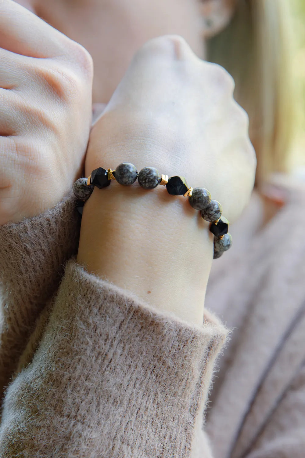 GŁĘBOKIE KORZENIE - BRANSOLETKA Z BRĄZOWEGO OBSYDIANU I ONYKSU na nadgarstku, z widocznymi okrągłymi kamieniami i geometrycznymi elementami, na tle delikatnej, beżowej tkaniny.