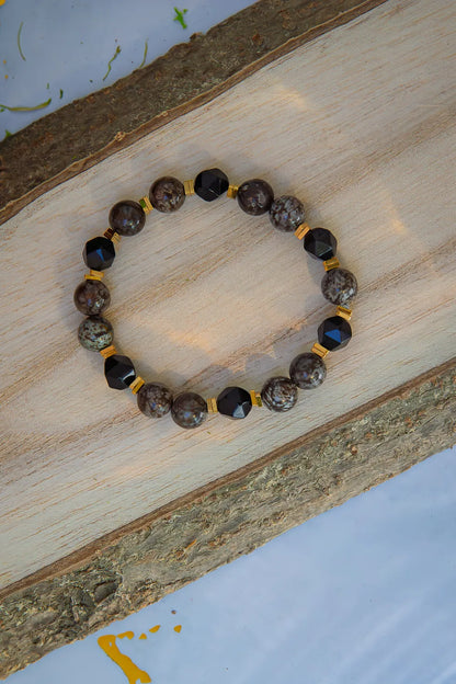 GŁĘBOKIE KORZENIE - BRANSOLETKA Z BRĄZOWEGO OBSYDIANU I ONYKSU, prezentująca się na naturalnym drewnianym tle. Biżuteria składa się z okrągłych kamieni onyksu i czarnych kryształów obsydianu, oddzielonych złotymi elementami.