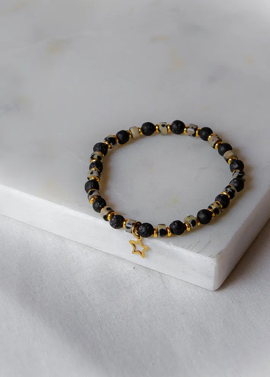 Gwiezdna Noc Bransoletka Czarna Lawy Złota Gwiazdka z kulkami z czarnej lawy, kamienia w cętki i złotymi elementami, na tle białego marmuru.