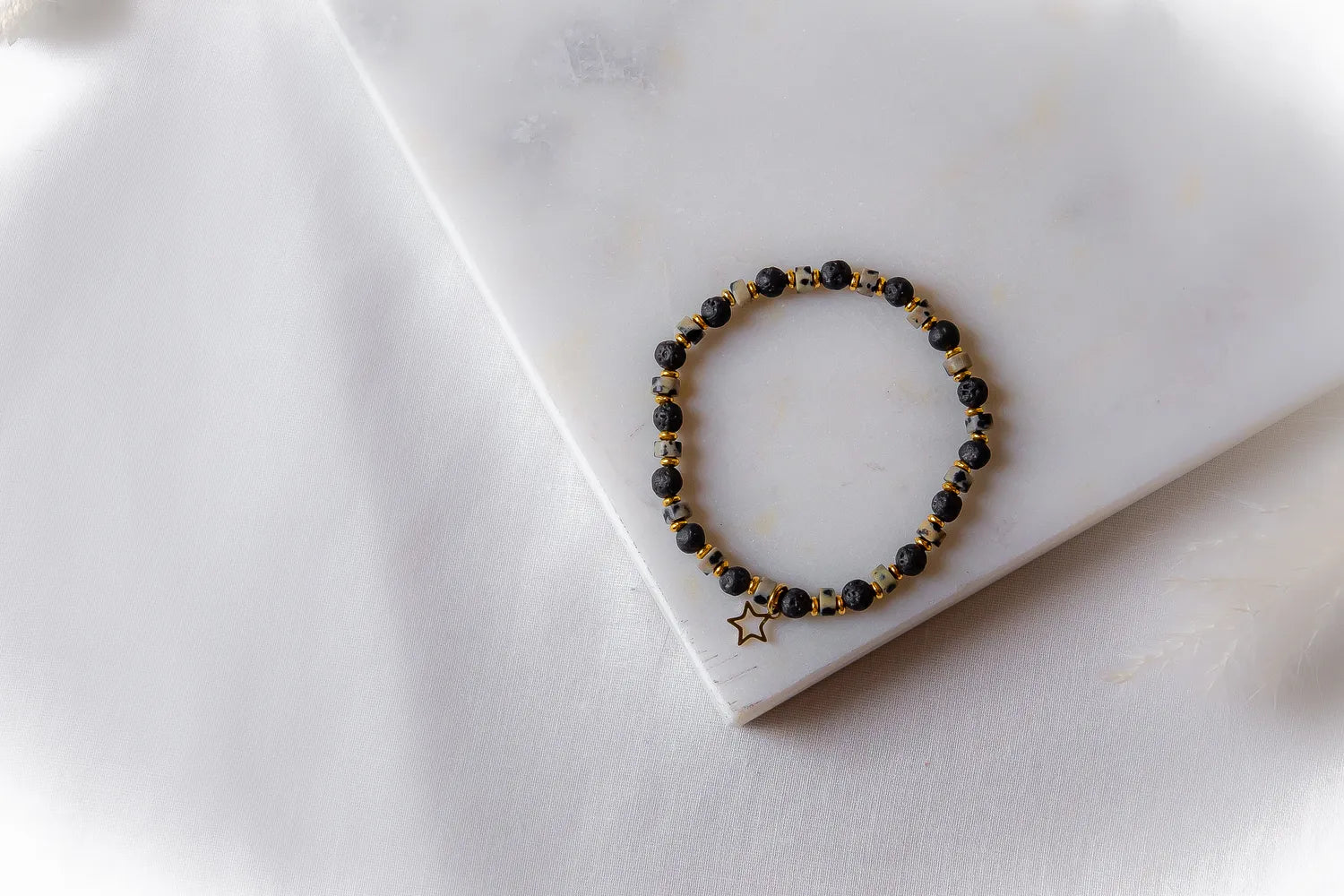 Gwiezdna Noc Bransoletka Czarne Kamienie z złotą zawieszką w kształcie gwiazdy, ułożona na białej marmurowej płycie.