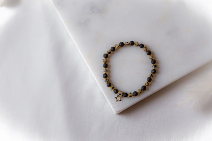 Gwiezdna Noc Bransoletka Czarne Kamienie z złotą zawieszką w kształcie gwiazdy, ułożona na białej marmurowej płycie.