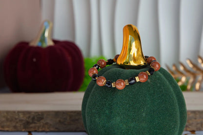 GWIEZDNA PUSTYNIA - BRANSOLETKA Z PIASKU PUSTYNI I CZARNEGO AGATU. Bransoletka z koralikami piasku pustyni i czarnego agatu, ułożona na zielonym podstawce w kształcie dyni, z pomarańczowym elementem na szczycie. Tło z czerwoną dynią i białymi zasłonami.