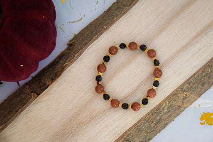 GWIEZDNA PUSTYNIA - BRANSOLETKA Z PIASKU PUSTYNI I CZARNEGO AGATU. Bransoletka z koralikami z piasku pustyni i czarnego agatu, ułożona na drewnianym tle, obok czerwonego kwiatu.