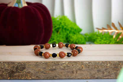 GWIEZDNA PUSTYNIA - BRANSOLETKA Z PIASKU PUSTYNI I CZARNEGO AGATU. Bransoletka z koralikami z piasku pustyni i czarnego agatu, ułożona na drewnianym bloku z zielonym tłem i dekoracją.