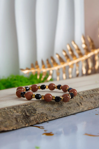 GWIEZDNA PUSTYNIA - BRANSOLETKA Z PIASKU PUSTYNI I CZARNEGO AGATU. Bransoletka z brązowymi koralikami piasku pustyni i czarnymi kamieniami agatu, ułożona na kamiennej powierzchni z tłem zielonej mchy i złotych liści.