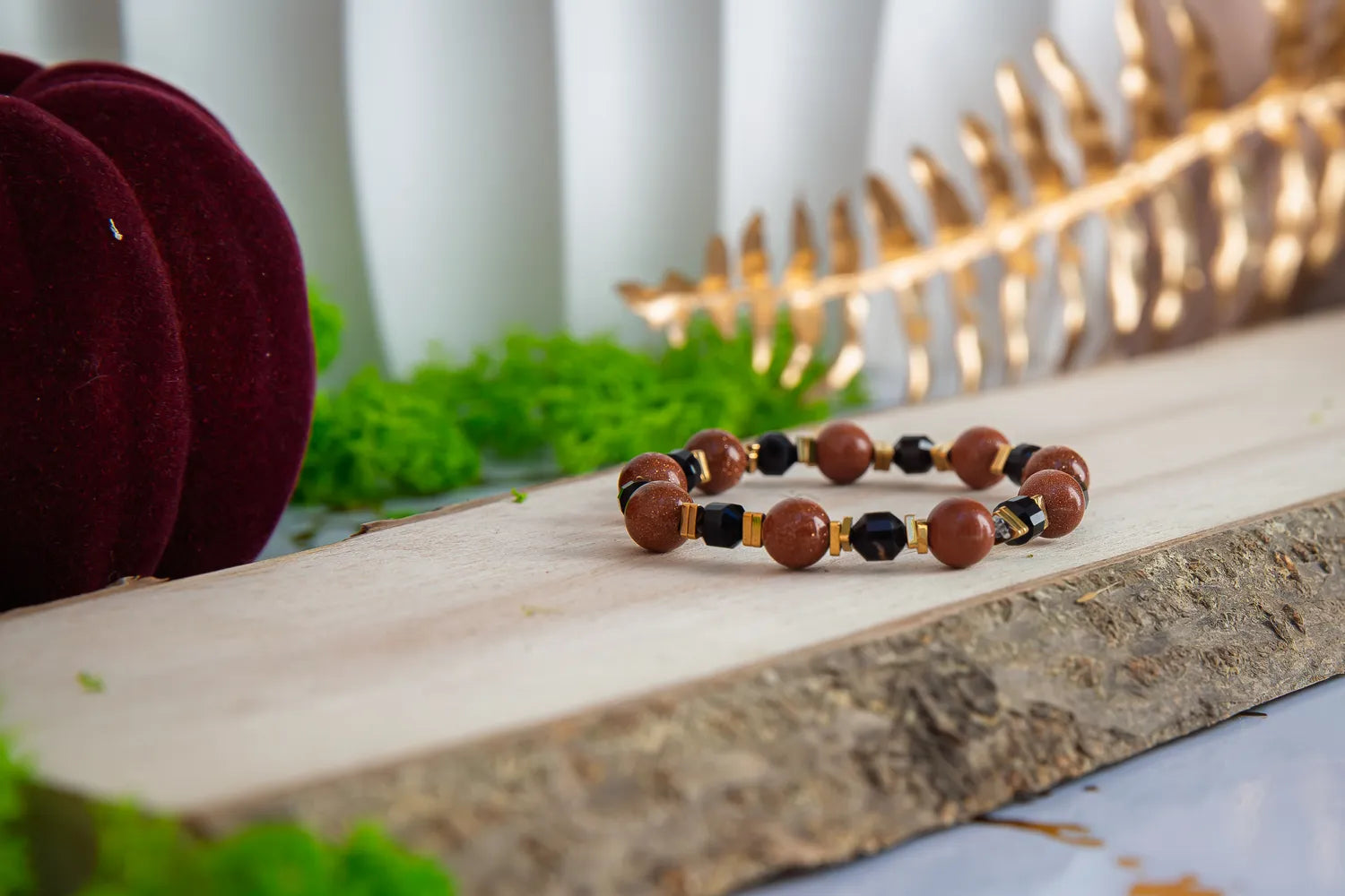 GWIEZDNA PUSTYNIA - BRANSOLETKA Z PIASKU PUSTYNI I CZARNEGO AGATU. Bransoletka z brązowymi i czarnymi koralikami leży na drewnianym pomoście, w tle zielone mchy i złota dekoracja.