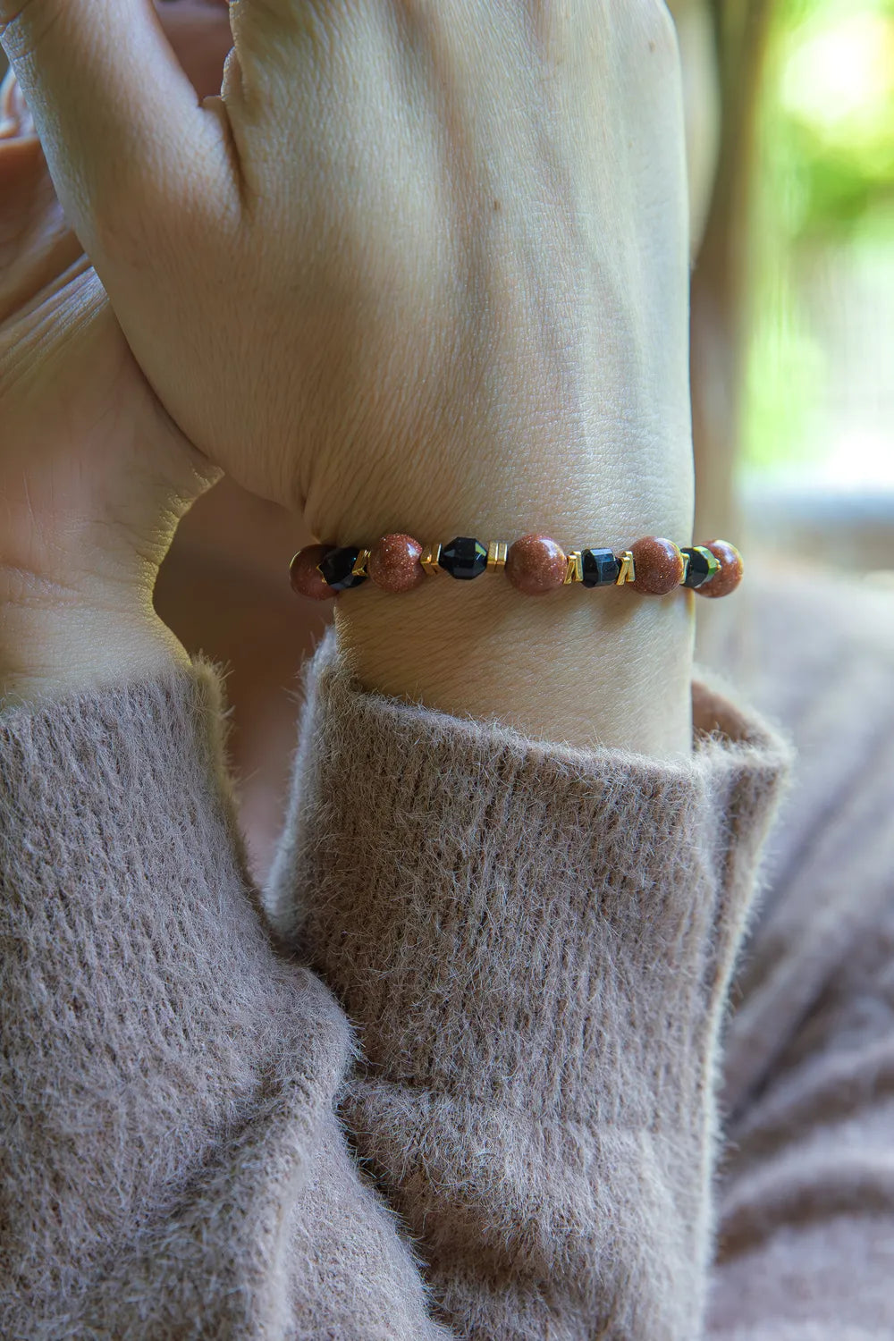 GWIEZDNA PUSTYNIA - BRANSOLETKA Z PIASKU PUSTYNI I CZARNEGO AGATU na nadgarstku, z widocznymi koralikami z piasku pustyni i czarnego agatu, na tle delikatnej, beżowej dzianiny.