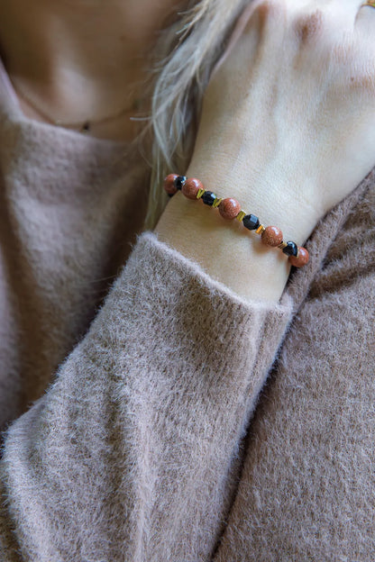 GWIEZDNA PUSTYNIA - BRANSOLETKA Z PIASKU PUSTYNI I CZARNEGO AGATU na nadgarstku, z widocznymi pomarańczowymi i czarnymi kulkami, na tle delikatnej, beżowej bluzy.