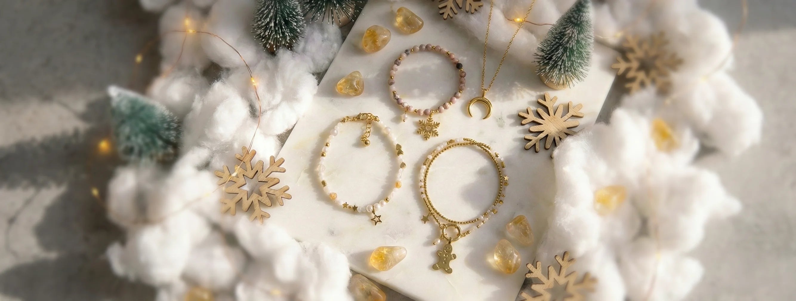 Świąteczna aranżacja biżuterii z bransoletkami z motywami piernikowego ludzika, gwiazdki i śnieżynki oraz naszyjnikiem z półksiężycem na marmurowej tacy. Dekoracja obejmuje mini choinki, bawełniany śnieg, drewniane śnieżynki i ciepłe oświetlenie LED.