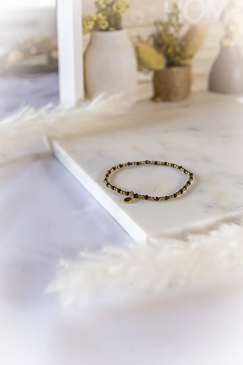 Klasyczna Elegancja Bransoletka Z Hematytu I Czarnego Turmalinu Marmur: delikatna, złota bransoletka z małymi, czarnymi kamieniami i owalnym medalionem na marmurowej powierzchni.