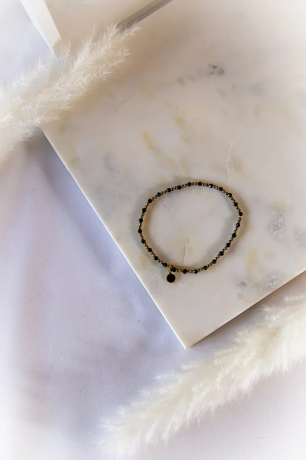 Klasyczna Elegancja Bransoletka Z Hematytu I Turmalinu - czarne, błyszczące kamienie na złotym druciku, z okrągłą czarną zawieszką, na marmurowej płytce.