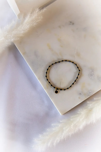 Klasyczna Elegancja Bransoletka Z Hematytu I Turmalinu - czarne, błyszczące kamienie na złotym druciku, z okrągłą czarną zawieszką, na marmurowej płytce.