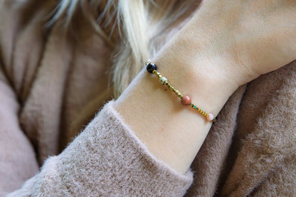 KOMPAS DUSZY - BRANSOLETKA Z TURMALINU, RODONITU I PIASKU PUSTYNI. Bransoletka z kolorowymi kamieniami na delikatnej ręce, tło z miękkiej, beżowej tkaniny.