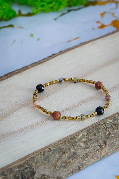 KOMPAS DUSZY - BRANSOLETKA Z TURMALINU, RODONITU I PIASKU PUSTYNI. Bransoletka z kolorowymi koralikami turmalinu, rodonitu i piasku pustyni leży na drewnianym tle, z naturalnym tłem wodnym w tle.