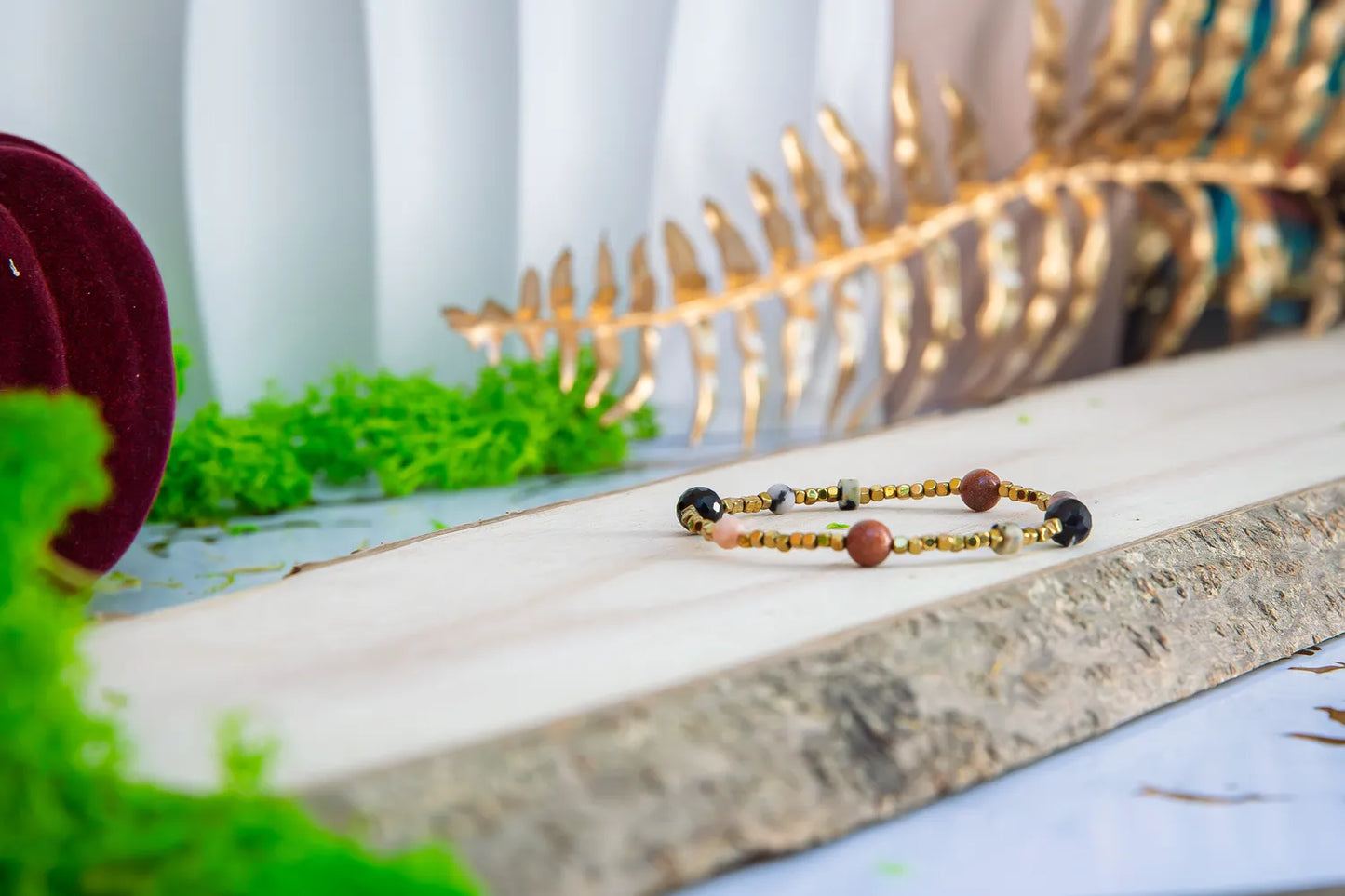 KOMPAS DUSZY - BRANSOLETKA Z TURMALINU, RODONITU I PIASKU PUSTYNI. Bransoletka z kolorowymi koralikami turmalinu, rodonitu i piasku pustyni leży na drewnianym tle, z zielonymi liśćmi i złotą dekoracją w tle.