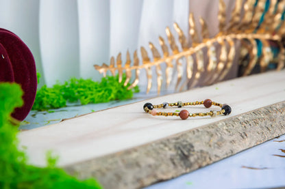 KOMPAS DUSZY - BRANSOLETKA Z TURMALINU, RODONITU I PIASKU PUSTYNI. Bransoletka z kolorowymi koralikami turmalinu, rodonitu i piasku pustyni leży na drewnianym tle, z zielonymi liśćmi i złotą dekoracją w tle.