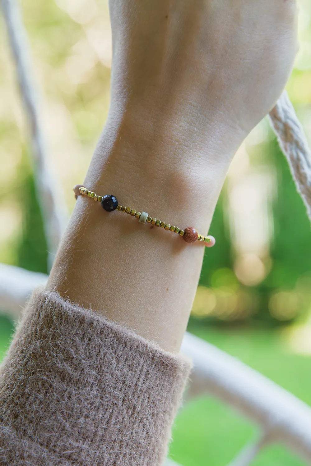 KOMPAS DUSZY - BRANSOLETKA Z TURMALINU, RODONITU I PIASKU PUSTYNI. Bransoletka z kolorowymi kamieniami na złotym łańcuszku, noszona na nadgarstku. Tło z zielonymi roślinami i drewnianym elementem.