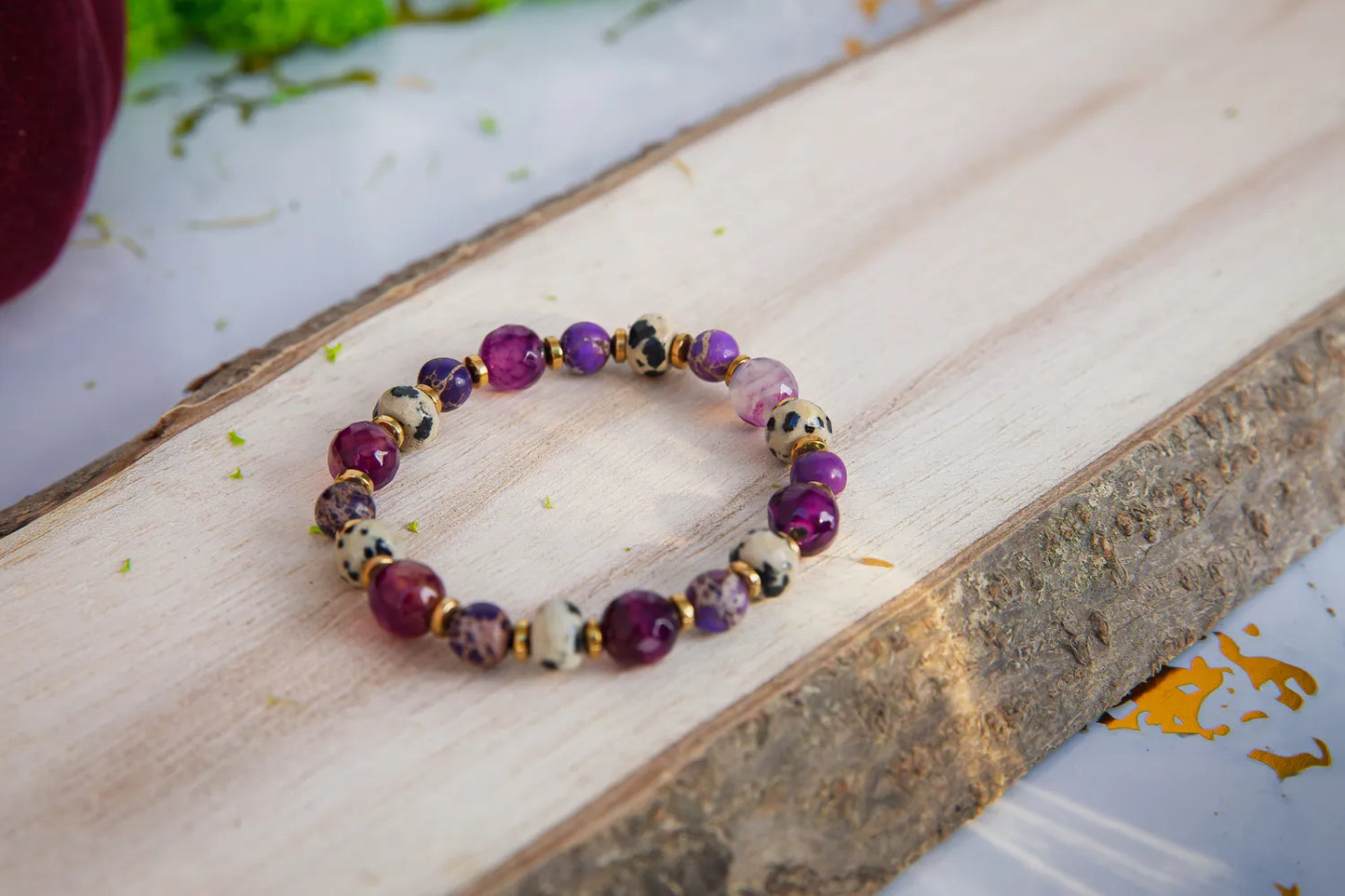 KRÓLEWSKA INTUICJA - BRANSOLETKA Z JASPISU CESARSKIEGO I FIOLETOWEGO AGATU. Bransoletka z kolorowymi kamieniami, w tym jaspisem cesarskim i fioletowym agatem, leży na drewnianym tle.
