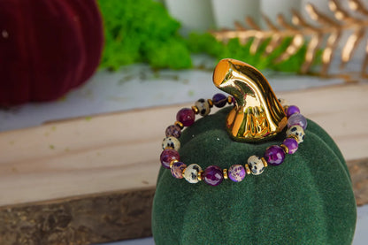KRÓLEWSKA INTUICJA - BRANSOLETKA Z JASPISU CESARSKIEGO I FIOLETOWEGO AGATU. Bransoletka z kolorowymi kamieniami, w tym jaspisem cesarskim i fioletowym agatem, na zielonym aksamitnym podstawce. Tło z dekoracjami roślinnymi i złotymi elementami.