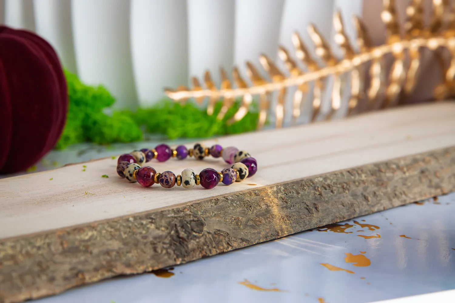KRÓLEWSKA INTUICJA - BRANSOLETKA Z JASPISU CESARSKIEGO I FIOLETOWEGO AGATU, prezentująca się na drewnianym pomoście z kamieniami w kolorach fioletu i brązu, na tle zielonych liści i złotych ozdób.