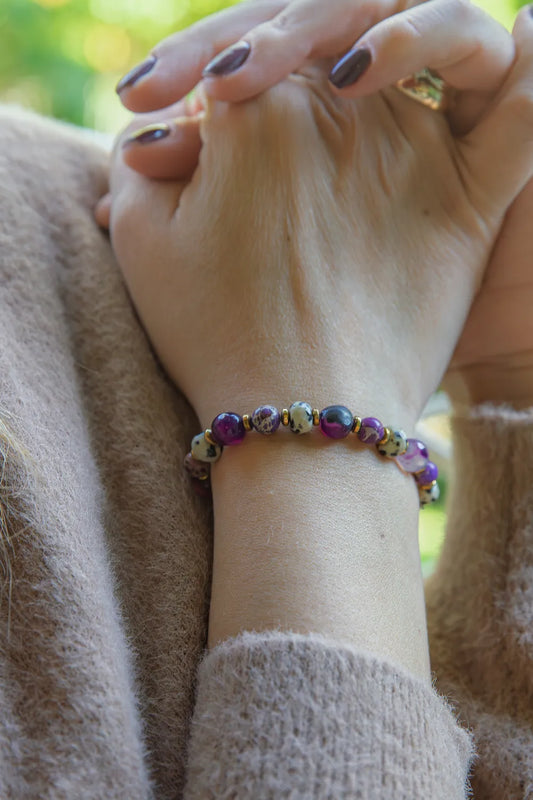 KRÓLEWSKA INTUICJA - BRANSOLETKA Z JASPISU CESARSKIEGO I FIOLETOWEGO AGATU na nadgarstku, z widocznymi kolorowymi kamieniami, na tle miękkiego, beżowego swetra.