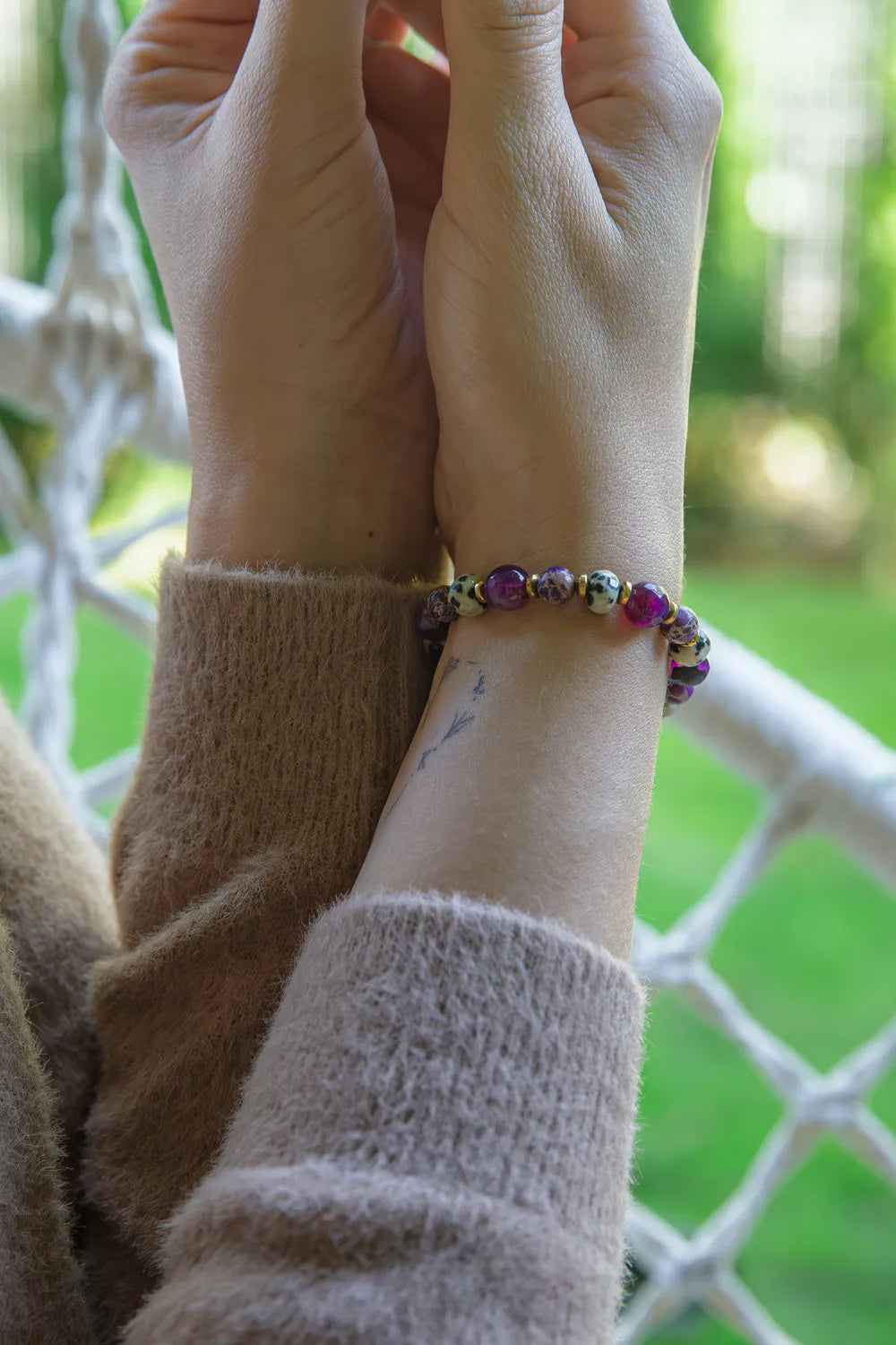 KRÓLEWSKA INTUICJA - BRANSOLETKA Z JASPISU CESARSKIEGO I FIOLETOWEGO AGATU na nadgarstku, z widocznymi kolorowymi kamieniami, na tle zielonej trawy i siatki.