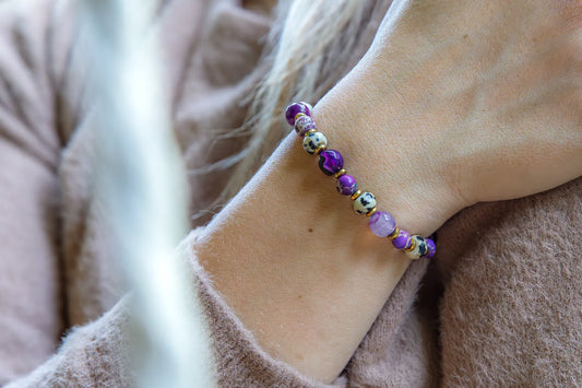 KRÓLEWSKA INTUICJA - BRANSOLETKA Z JASPISU CESARSKIEGO I FIOLETOWEGO AGATU. Bransoletka z kolorowymi kamieniami, w tym jaspisem cesarskim i fioletowym agatem, na tle delikatnej, beżowej tkaniny.